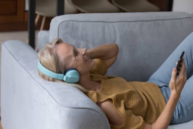 Büyük kablosuz kulaklıklı mutlu orta yaşlı kadın cep telefonuyla online oyuncunun rahatlatıcı müziğini dinliyor, akıllı telefonda medya uygulaması kullanıyor, kanepeye uzanmış, gözleri kapalı gülüyor.