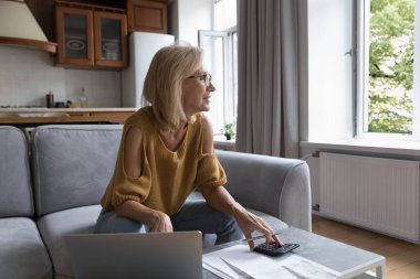 Mutlu ev sahibi kadın aylık evrak işleri, gelir, planlama tasarrufları, bütçe, evde finans yönetimi işi, dizüstü bilgisayar, hesap makinesi, pencereden dışarı bakarak düşünüyor.