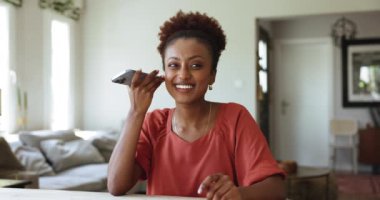 Afrikalı bir kadın oturma odasında oturmuş cep telefonundaki sesli mesajları dinliyor, arkadaşa cevap gönderiyor, sesli mesajları kaydediyor, modern akıllı telefonu kullanarak hoparlörden konuşuyor. Teknoloji, uzaktan iletişim.
