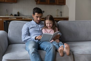 Sevecen baba ve tatlı anaokulu kızı birlikte kitap okuyorlar, sarılıyorlar, kanepede oturuyorlar, evde birlikte vakit geçiriyorlar. Yakışıklı baba neşeli çocuğa masal anlatıyor.