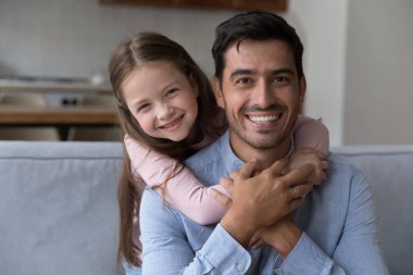 Neşeli şirin anaokulu kızı babaya kanepede sarılıyor, kameraya bakıyor, gülümsüyor, gülüyor. Mutlu baba, küçük kızı sırtında taşıyor, evde çocukla oynuyor. Kafasından vurulmuş.
