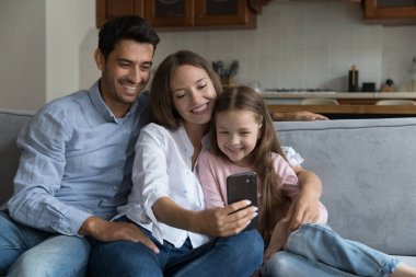 Mutlu bir çift ebeveyn ve mutlu bir kız çocuğu video görüşmesinde konuşuyor, cep telefonuyla selfie çekiyor, sarılıyor, kanepede birlikte oturuyor, gülüyor, internet uygulaması kullanıyor, eğleniyor.