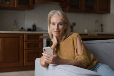 Olumlu tatmin olmuş akıllı telefon kullanıcısı kadın portresi. Orta yaşlı kadın elinde akıllı telefon tutuyor, internet uygulaması kullanıyor, servis yapıyor, kanepeye cihaz dayamış, kameraya bakıyor.