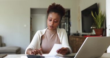 Afrikalı bir kadın dizüstü bilgisayarla masasında oturuyor. Masa başında harcamalar ve gelir hesaplamaları yapıyor, aile harcamalarını gözden geçiriyor, bütçe hesaplamalarını yapıyor, mali verileri analiz ediyor, muhasebe işlerini ev ofisinden yapıyor. Mali yönetim