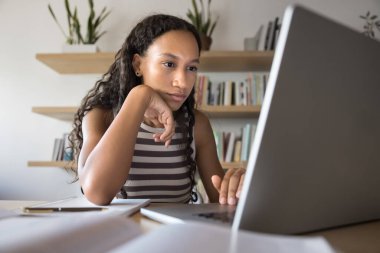 Düşünceli genç Afrikalı öğrenci kütüphanede dizüstü bilgisayarda makale okuyor. Sınıf raporunu düşünüyor, makale yazıyor, çeneye dokunuyor, araştırma kağıdı için internette geziniyor.