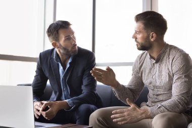 Two serious male business partners talking at laptop, sitting on office coach, discussing teamwork, work project. Consultant giving advice, to client, manager, entrepreneur, businessman clipart