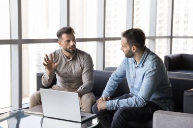 Serious busy professional man talking to business colleague, moving hands for explaining. Two entrepreneurs men meeting in office co-working hall, sitting on sofa, discussing teamwork, collaboration clipart