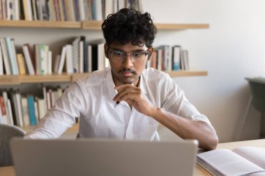 Gözlüklü yakışıklı Hintli öğrenci dizüstü bilgisayarda ders çalışıyor, konferans izliyor, Webinar, sınıf, kütüphanede video dersi öğreniyor, internet teknolojisi kullanıyor, araştırma ödevi yapıyor.