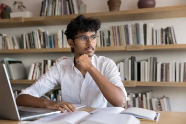 Gözlüklü zeki Hintli öğrenci bilimsel makale yazıyor, kütüphanede araştırma kağıdı yazıyor, dizüstü bilgisayarda oturuyor, açık kitap okuyor, çeneye dokunuyor, başka yere bakıyor, kompozisyon üzerinde düşünüyor, rapor öğreniyor.