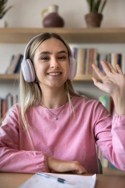 Kulaklıklı mutlu genç kız öğrenci kameraya el sallıyor, gülümsüyor, konferans veriyor, internette konuşuyor, gülümsüyor. Dikey kafa resmi