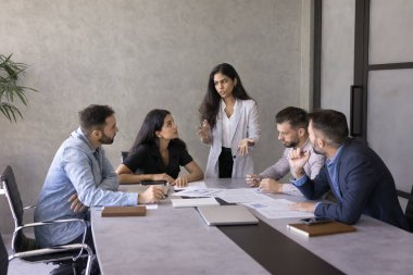 Genç Hintli iş projesi yöneticisi kadın pazarlama raporlarını sunuyor, satış verilerini ekibe sunuyor, iş arkadaşlarıyla konuşuyor, büyük toplantı masasında duruyor. Finansal araştırmaları analiz eden çeşitli grup