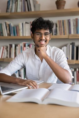 Gözlüklü mutlu Hintli öğrenci kütüphanede arka planda kitaplıklarla poz veriyor, açık kitaplarda oturuyor, çeneye dokunuyor, kameraya bakıyor, gülümsüyor. Dikey çekim