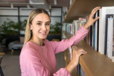 Pozitif genç kız öğrenci kitaplıktan ders kitabı seçiyor, üniversite kütüphanesini ziyaret ediyor, kameraya bakıyor, gülümsüyor, bilgi almak için kitap alıyor, kitapçıdaki kitapçılarda portre için poz veriyor.