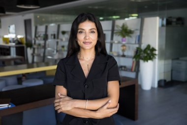 Kendine güvenen, ortadoğulu iş kadını modern çalışma ortamında portre için poz veren, elleri güven jesti ile katlanmış kameraya bakan. Genç Arap iş kadını kafasından vurulmuş.