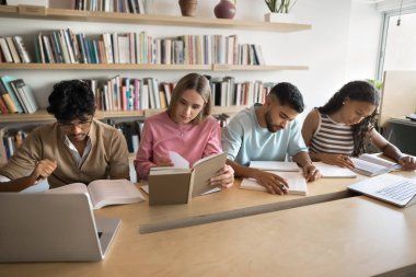 Çok ırklı üniversite öğrencileri kütüphaneyi ziyaret ediyor, ders kitapları okuyor, notlar yazıyor, proje raporlarını hazırlıyor, öğrenim kitapları okuyor, birlikte büyük bir masada oturuyor, sessiz kalıyorlar.