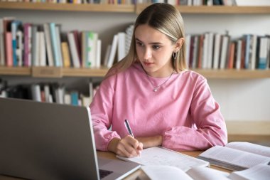 Odaklanmış zeki kız öğrenci kütüphanede dizüstü bilgisayardan Webinar öğrenmeyi izliyor, özet yazıyor, notlar yazıyor, sınava hazırlanıyor, internetten öğrenim görüyor, internet teknolojisini eğitim için kullanıyor.