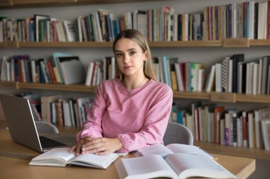 Gerçekten güzel bir beyaz kız öğrenci lise kütüphanesinde üniversite sınavına hazırlanıyor, bir yığın kitap ve dizüstü bilgisayarla masada oturuyor, fotoğraf için kameraya bakıyor.
