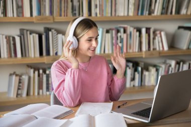 Kablosuz kulaklıklarla el sallayan, online öğrenim konferansında konuşan, video konuşmasında konuşan, kütüphanede kitaplarla masada oturan mutlu, zeki, genç üniversite öğrencisi kız.