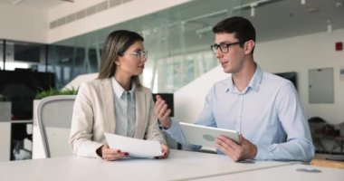 Modern ofis ortamında masa başında oturan, elinde kağıtlar tutan, tablet kullanan, takım çalışmasına katılan, kağıtları inceleyen, mali belgeleri veya bütçeyi tartışan, müşterilere sunum hazırlayan iki meslektaş