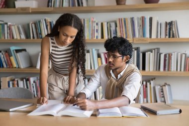 Çeşitli zeki genç öğrenciler halk kütüphanesinde üniversite ödevi yapıyorlar, birlikte kitap okuyorlar, sayfayı işaret ediyorlar, araştırma çalışmasını tartışıyorlar, öğrenme projesini tartışıyorlar, sınıfa rapor veriyorlar.