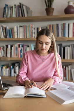 Kütüphanedeki masada ders kitabı okuyan, makale, makale, araştırma çalışması, sınıf raporu için notlar yazan, eğitim sınavına hazırlanan, odaklanmış genç bir üniversite öğrencisi. Dikey çekim