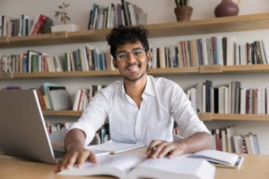 Gözlüklü neşeli Hintli genç kolej öğrencisi halk kütüphanesinde bir yığın kitapla oturuyor, üniversite sınavına hazırlanıyor, kameraya bakıyor, gülümsüyor, portre için poz veriyor.