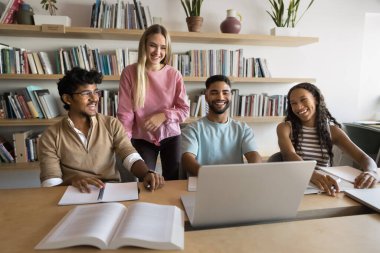Mutlu öğrenci kızlar ve erkekler kütüphanede toplanıyor, dizüstü bilgisayarları açık bir masada oturuyor, video izleyip araştırma çalışmalarını tartışıyor, gülüyor, üniversite arkadaşlığından keyif alıyorlar.