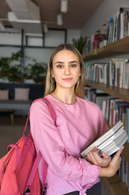 Kitap raflarında yığınla kitap tutan, kameraya bakan, gülümseyen, yüksek öğrenim için bilgi toplayan, devlet üniversitesi kütüphanesinde, kitapçıda poz veren kendinden emin bir öğrenci. Dikey çekim