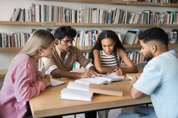 Çok ırklı çalışkan öğrencilerden oluşan bir grup kütüphanede ders kitabı okuyor, bilimsel makaleler okuyor ve tartışıyor, kalemleri sayfaya doğrultuyor, grup raporu üzerinde çalışıyor, araştırma makalesi üzerinde çalışıyor.