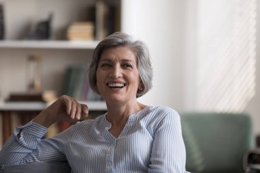 Çekici orta yaşlı kadın oturma odasındaki rahat koltukta dinleniyor kameraya bakıyor huzurlu görünüyor, mutlu hissediyor, evde tek başına vakit geçiriyor. Ev sahibi portresi, emeklilik konseptinde tasasız bir hayat.