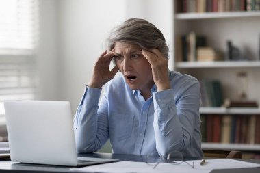 Stressed aged businesswoman staring at laptop hold head feels shocked by bad news, unsaved project or information, financial problem, work-related mistake, company bankruptcy, business failure concept clipart