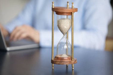 Masadaki klasik kum saati, iş kadını ya da ofis çalışanı iş yerinde, arka planda oturan dizüstü bilgisayarla çalışıyor. Acele et, teslim tarihi, verimlilik ve verimlilik kavramı.