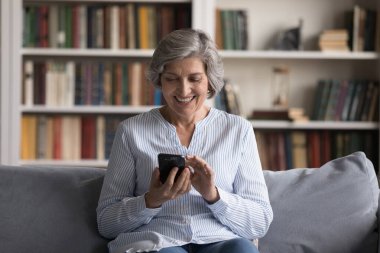 Olgun 60 'lı yaşlardaki kadınlar koltukta oturup gülümser cep telefonu ekranına bakar, mesaj gönderir, kurye kullanır, yetişkin çocuklarla sosyal medyada sohbet eder, uzaktan iletişimin keyfini çıkarır, modern teknoloji ve internet kavramını kullanır.