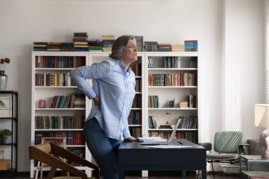 60 'ların iş kadını sert sırt kaslarına dokunuyor, ani ağrılar çekiyor, ofiste masanın yanında duruyor, güçlü bel ağrısı sağlıksız görünüyor. Sağlık sorunu, yanlış duruş, sabit çalışma romatizması konsepti