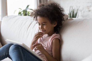 Küçük Afrikalı çocuk tablet bilgisayarda çizgi film izliyor, online kitap okuyor, okul ödevi yapıyor, kanepede boş vakitlerin tadını çıkarıyor, çalışmaktan zevk alıyor, video oyunu oynuyor.