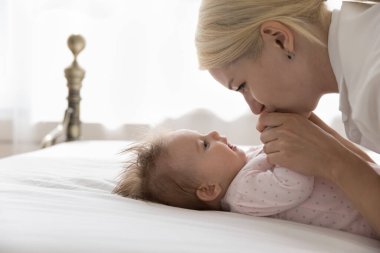 Mutlu, genç, güzel bir anne kucağında tatlı bir bebekle yatağa uzanıyor, kucağında bebek kollarıyla öpüyor. Sadık anne küçük çocukla sevgi, şefkat, neşe, annelik keyfi, aile zamanı ile oynuyor.