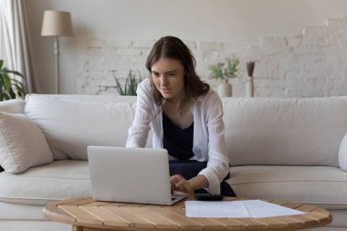 Odaklanmış genç kiracı kız evde evrak işleriyle uğraşıyor, dizüstü bilgisayarda online ödeme uygulamasını kullanıyor, internete giriyor, kağıtları kontrol ediyor, faturaları ödüyor, kira ücretlerini ödüyor, vergileri ödüyor, bütçe sayıyor.