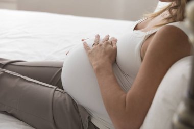 Hamile kadın beyaz kolsuz tişörtün altında 9 aylık bir tümseğe dokunuyor, bebek tekmelerini hissediyor. Annemin yatakta dinlenmesini, dinlenmesini evde eğlenmesini, karnını kucaklamasını bekliyorum. Kesilmiş atış. Hamilelik konsepti