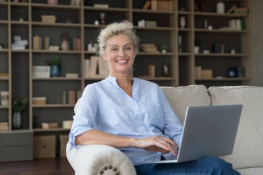 Neşeli, güzel, son sınıf serbest kadın ev portresi. Serbest çalışan mutlu bir iş kadını, dizüstü bilgisayarı olan bir girişimci, dairede koltukta oturuyor, dişlek gülümsemeli kameraya bakıyor.