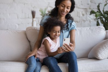 Mutlu Afrikalı anne ve neşeli genç kız akıllı telefondan video çekiyor, selfie çekiyor, cep telefonundan alışveriş yapıyor, cep telefonundan öğrenme uygulamasını kullanıyor, evde dinleniyor.