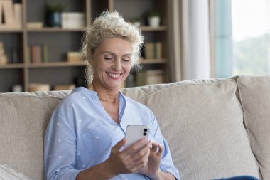 Mutlu olgun bir kadın evde online başvuru kullanıyor, cep telefonu tutuyor, kanepede dinleniyor, boş vakitlerin tadını çıkarıyor, internet iletişiminin, kablosuz teknolojinin, gülümsüyor, gülüyor.