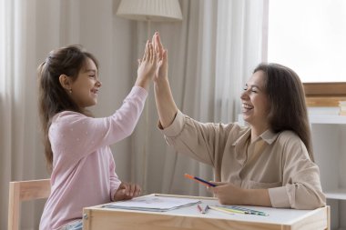 Neşeli anne ve pozitif anaokulu çocuğu oyun odasında birlikte resim çiziyor, renkli çizimlerle kağıt üzerinde beşlik çakıyor, kızları kutluyor, sanatsal eğitim veriyor, gülümsüyor, gülüyor.