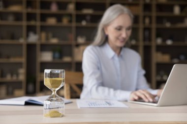 Kum camı, yaşlı iş kadınının çalışma masasına yakın planda bulanık dizüstü bilgisayar kullanarak. Yetişkin profesyonel takip programı, son teslim tarihinden önce proje görevlerini tamamlamak. Zaman yönetimi kavramı