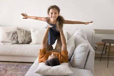 Güçlü bir baba küçük kızını havaya kaldırıyor, sırt üstü uzanıyor, çocuğuyla uçuyor. Mutlu tatlı küçük kız kameraya açık elleriyle bakıyor, babasıyla aktif bir oyun oynuyor.