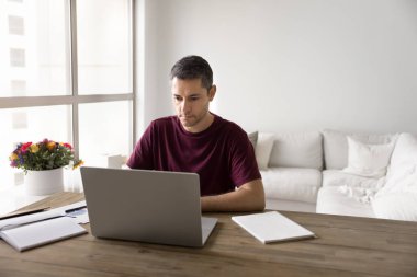 Odaklanmış orta yaşlı, serbest çalışan, sıradan tişörtlü İspanyol bir iş adamı evde online proje üzerinde çalışıyor. iş yerinde dizüstü bilgisayarda yazı yazan ciddi girişimci, iş için internet teknolojisini kullanıyor