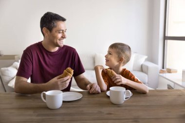 Mutlu İspanyol baba ve pozitif tatlı çocuk masada sıcak içeceklerle kruvasan yiyorlar, rahat bir evde kahvaltı yapıyorlar, tatlı atıştırmalıkların tadını çıkarıyorlar, konuşup gülüyorlar.