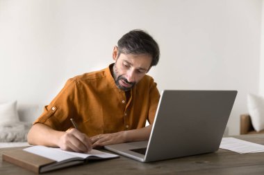 Odaklanmış orta yaşlı Latin girişimci, dizüstü bilgisayarda çalışıyor, not defterine notlar yazıyor, video konferans görüşmesi yapıyor, profesyonel Webinar 'ı izliyor.