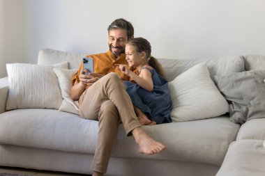 Küçük tatlı bir kız çocuğu ve orta yaşlı İspanyol bir baba cep telefonunu evdeki kanepede kullanıyor ve internet iletişiminin tadını çıkarıyor. Neşeli baba kızına online alışveriş uygulaması gösteriyor