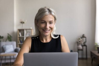 Melez iletişimi olan gülümseyen yaşlı bayan evdeki bilgisayarı kullanarak konferans uygulaması sohbetinde görüntülü konuşma yaptı. Mutlu olgun kadın kendine güveni olan bilgisayar kullanıcısı masa başında oturup kitap okumanın keyfini çıkar.