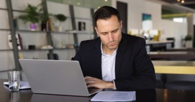 Ciddi Avrupalı yetişkin işadamı iş meselelerini uzaktan çözer, masa başında dizüstü bilgisayar kullanır, organizatöründe internetten bulunan bilgileri yazar, planlama işi, rapora odaklanmış görünür.